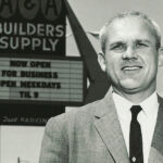 Black & White photo of Buzz Oates in a suite in front of A&A Builder's Supply sign